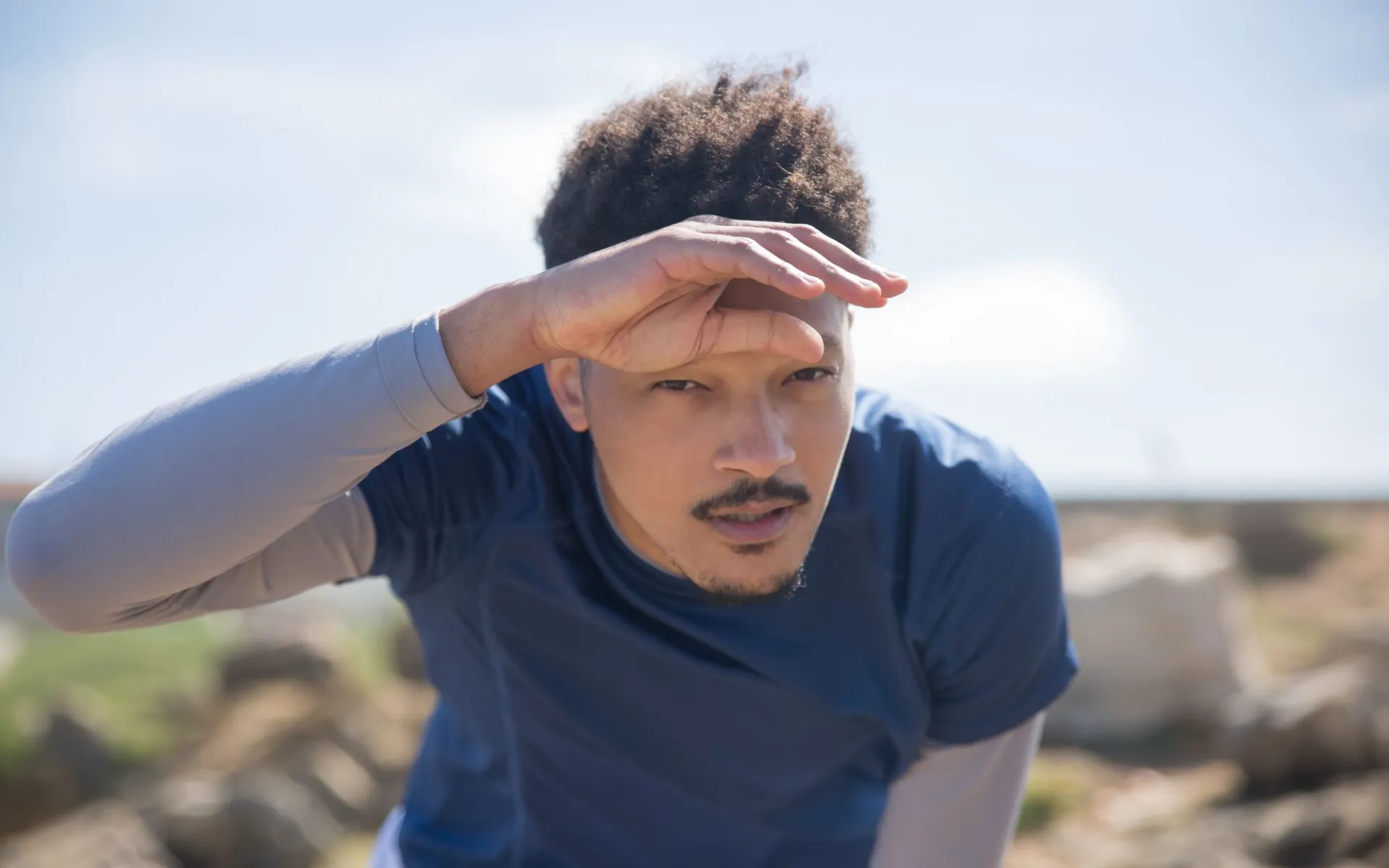 Homem com camiseta azul fazendo sombra nos olhos com a mão em um dia de sol forte ao ar livre